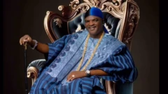 Obesere jòkòó lórí àga, wọ aṣọ olómi aro, mú ọ̀pá dání, wọ ṣéìnì sọ́rùn 