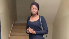 Noella Mukumbi, de pé em frente a uma escada, com blusa e calça preta e carregando uma bolsa da mesma cor, sorri para a câmera