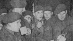 A grainy black-and-white photograph from a newspaper showing paratroopers gathered in a group drinking tea. Each is wearing their beret. It shows a moment just before they got into planes to be dropped over German lines at the River Rhine in Operation Varsity, March 1945.