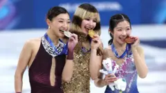 3人がリンクで横並びに立ち、左手で持ったメダルを歯でかむようなポーズをとっている。中井の右手には大会マスコットのぬいぐるみが握られている