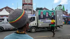 Caravana de campaña del APNU. Un camión pasa con un cartel que anuncia a varios candidatos del partido. Un hombre de espaldas observa el vehículo; viste una camisa azul a rayas y un gorro rastafari.