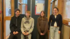 Vier Teenager standen lächelnd auf dem Schulkorridor – zwei Jungen und zwei Mädchen. Sie tragen Freizeitkleidung.