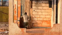 Umugabo upfukamye ku mavi mu nyubako y'amatafari arebera mu idirishya. 