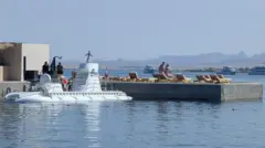 A submarine on a dock in harbour next to tourist deck chairs. සංචාරකයින් වාඩිවී සිටින පුටු අසල වූ නැව් තටාකයක නැංගුරම්ලා ඇති සබ්මැරීනයක්. 