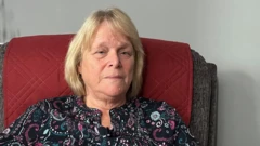 Ifoto y'umugore usa n'unaniwe, wambaye ishati y'amabara menshi, aho yicaye mu ntebe yegamye