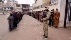 صورة لصفين من عشرات الرجال الذين يرتدون معاطف شتوية أمام أحد المباني، ويقف رجل بزي عسكري حاملاً سلاحاً أمام الصفين.