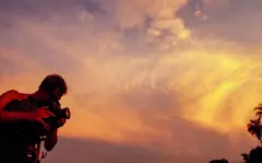 A imagem mostra uma pessoa operando uma câmera de vídeo profissional ao ar livre durante o pôr do sol. Em silhueta contra um céu alaranjado e repleto de nuvens iluminadas, o cinegrafista aparece concentrado na filmagem, enquanto a luz quente do entardecer domina a composição. Ao fundo, a copa de uma palmeira reforça a atmosfera tropical da cena.