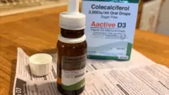 Frasco de vitamina D3 em gotas para uso oral, com sua embalagem e bula sobre uma mesa