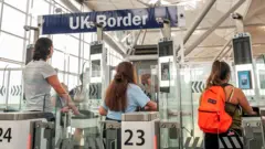 People approach electronic passport scanners at the UK border at Stansted airport in 2018 - one person wears an orange backpack and all have their backs to the camera. In the background is a sign saying 'UK Border'.