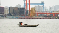 Homem de pé em uma minúscula embarcação na baía de Dalian, na China, tendo ao fundo o estaleiro com enormes guindastes e outras máquinas. Uma grande estrutura azul traz o nome da empresa Dalian Shipbuilding Industry Co. Ltd.