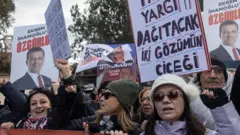 Üç kadın bir pankart tutuyot ikisi güneş gözlüklü ve bereki biri ekrana bakarak gülümsüyor. Geride pankartlar ve imamoğluna özgürlük sloganlı afişler var. 