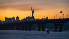 Зимовий краєвид Києва під час заходу сонця. На передньому плані – вкрита льодом поверхня Дніпра, по якій ідуть двоє людей. Над ними проходить міст, уздовж якого тягнеться лінія ліхтарів. На горизонті видно темний силует міських будівель та велику фігуру Батьківщини-матері.