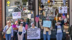 Um pequeno grupo de manifestantes se reúne em frente a uma loja. Elas carregam bandeiras americanas e cartazes com os dizeres "Se você não está preocupado com o nosso país, você não está prestando atenção" e "Este é o governo com o qual nossos fundadores se preocupavam".