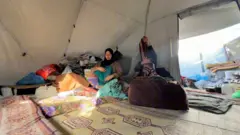 Amani Abu Salmiya, usando um lenço na cabeça, está sentada em sua tenda em meio aos restos de seu enxoval de casamento, devorados por roedores.