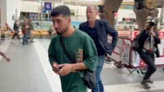 شاب ملتحٍ أدار وجهه للصحفي أندرو هاردينج متجهاً إلى مخرج محطة برمنغهام نيو ستريت. تظهر في الخلفية أرجل تمثال الثور الميكانيكي، ويظهر في الصورة أيضاً مصور من بي بي سي.