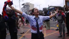 A young woman, wearing what resembles a school uniform which has been emblazoned with protest slogans, shouts and holds her arms and the Nepali flag in the air