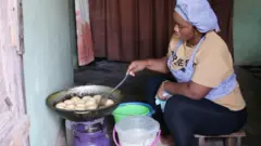 Oluwatomisin Onipuffpuff joko lori apoti ninu yara ka, o n din puffpuff lori gaasi idana. O wọ asọ ileke alawọ buluuu lori bulaosi alawọn paali, o si wọ sokoto alawọ dudu. Awọn ike meji ti eroja Puffpuff to n din wa niwaju rẹ, to si n fi sibi yi awọn puffpuff to n din pada.