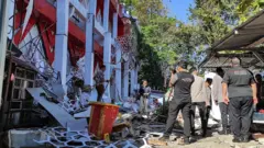 Solda kısmen yıkılmış bir bina, sağda 5-6 yetkili. Havanın güneşli, ılık olduğu anlaşılıyor. 