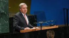 António Guterres ahagaze imbere y'indangururamajwi ebyiri, ziri ku meza maremare yo kuvugiraho ijambo, ariho ikirango cy'Umuryango w'Abibumbye.