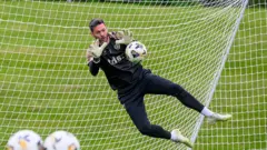 Craig Gordon faz uma defesa durante um treinamento pelo seu clube, o Heart of Midlothian, em Edimburgo, na Escócia, vestido de preto, pulando com as mãos abertas para impedir o gol