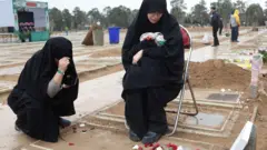 墓の前で黒い布で全身を覆った1人がしゃがみ、もう1人がパイプ椅子に腰かけて下を見ている。少し離れたところに他の人たちもいる