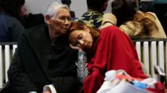 A woman covered in a red blanket rests with her eyes closed on the shoulder of an elderly lady in a black coat. Both are sitting on a white bench with crowds of others in the background. 