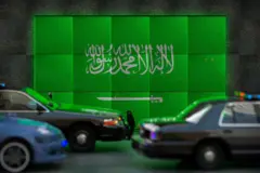 Carros passando em frente a tela com bandeira da Arábia Saudita