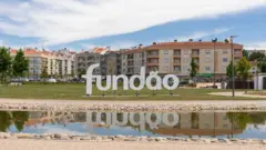 Um letreiro com o nome da cidade Fundão, em frente a edifícios baixo e com um curso de água em primeiro plano