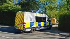 Three police vans in a row, parked in a a small side road. There are trees either side of the road.