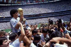 El futbolista Diego Maradona con el trofeo de FIFA en sus manos, cargado por personas en el campo de fútbol celebrando. En el fondo se ven las graderías repletas de gente