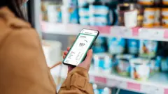 Osoba o niewidocznej twarzy stoi przed półką sklepową z nabiałem. W dłoni trzyma smartfon z aplikacją pokazującą wartość kaloryczną produktu. 