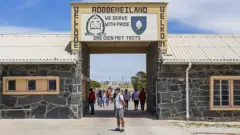 Turista em frente à porta da prisão de Robben Island, na África do Sul, com seus dizeres em inglês e afrikaans.