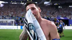 Lionel Messi en un estadio de fútbol, junto al campo, secándose las lágrimas del rostro con una camiseta mientras lleva un brazalete azul de la FIFA.