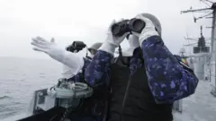 フリゲート艦の甲板で兵士2人が双眼鏡を手に監視している