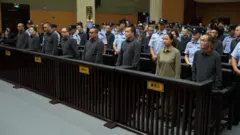 Filas de convictos chinos vestidos con camisetas y pantalones oscuros de manga larga se encuentran en el banquillo de los acusados ​​en un tribunal chino, flanqueados por guardias de seguridad uniformados.