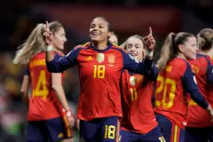 Vicky López, vestida con el uniforme de la selección española, sonríe mientras sus dedos índices apuntan hacia el cielo. En el fondo se ven otras futbolistas españolas