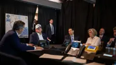 O presidente Donald Trump está sentado, usando um boné branco com a inscrição "USA", enquanto ouve o diretor da Agência Central de Inteligência (CIA), John Ratcliffe, falar. Ao redor da mesa estão o secretário de Estado dos EUA, Marco Rubio, e a chefe de gabinete da Casa Branca, Susie Wiles.