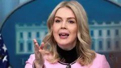 ピンクのジャケットを着たレヴィット氏が、ホワイトハウス会見場で記者団に向かって、右手を差し出して話している。。近くには米国旗がある