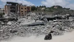 空爆により街の一区画が広範囲に破壊され、建物のがれきが積みあがっている。その前の道路を、男性2人が乗ったバイクが通っている