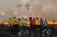 Grupo de pessoas usando roupas de proteçaõ amarela. Atrás há um incêndio