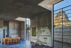Interior de arquitectura de hormigón visto con mesa de madera y grandes ventanales que enmarcan un paisaje rocoso.