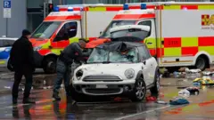 Two emergency service workers dey inspect one damaged white mini cooper.
