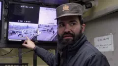 Um homem de barba apontando para um monitor de vigilância