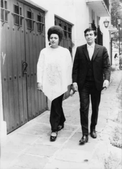 Alejandra Jáidar, al lado izquierdo, camina junto a Edmundo de Alba en la calle. Ella lleva el cabello recogido y un huipil blanco y un pantalón oscuro. Él va de traje oscuro, camisa y corbatas del color claro