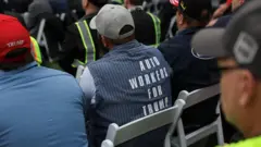 Um homem vestindo uma jaqueta com a inscrição "Auto Workers For Trump" está sentado no Jardim das Rosas durante o discurso de Trump na noite de quarta-feira. Ele também usa um boné de beisebol e está acompanhado por outros homens com roupas de trabalho.