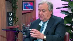 Antonio Guterres gestures as he talks into a microphone with a plant in background during an interview with BBC