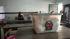 একটি টেবিলের ওপর ব্যালট ভর্তি ভোটের বাক্স রাখা। পেছনে আরেকটি বেঞ্চের ওপর একই ধরনের আরেকটি বাক্স। তার পেছনে একটি বেঞ্চে বসে আছেন একজন নারী