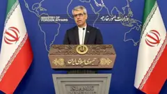 بقايي پر دریځ ولاړ دی، خبرې کوي او دواړو خواو ته یې د ایران بېرغونه ولاړ دي.