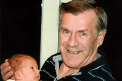 Fotografia de Ronnie Lockwood (à direita) sorrindo enquanto segura um bebê. Ele tem olhos azuis brilhantes e sorri para a câmera. Sua mão esquerda segura cuidadosamente a cabeça do bebê.