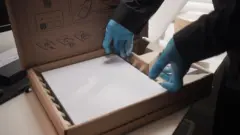 جهاز ستارلينك أبيض مستطيل الشكل داخل صندوق من الورق المقوى البني. تلمسه يدا شخص يرتدي قفازات بلاستيكية زرقاء رقيقة وملابس داكنة من الحواف، كما لو كان يرفعه من الصندوق.